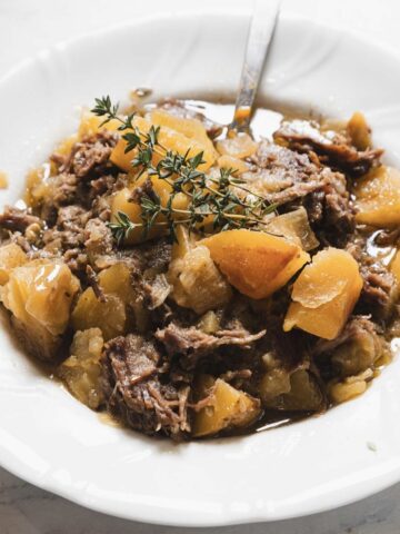 A white plate holds a savory Maple-Dijon Pot Roast stew with tender chunks of meat, potatoes, and vegetables in a rich brown broth. A sprig of fresh thyme garnishes the dish, adding a touch of elegance. A spoon rests in the bowl, inviting you to savor every bite.