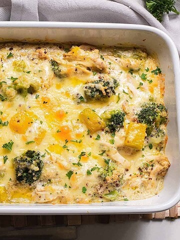 A baked casserole dish featuring quinoa, tender chicken, broccoli, and pumpkin chunks is topped with melted cheese and garnished with chopped herbs. The healthy meal rests on a woven mat, partially draped by a gray cloth.