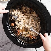 Two hands use forks to shred tender Greek chicken inside a black slow cooker. The dish brims with vegetables and herbs, offering a delightful aroma. A small bowl of seasonings sits invitingly in the background on a marble countertop.