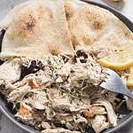 A plate featuring Greek chicken is loaded with shredded meat mixed with chopped vegetables and olives, garnished with a sprig of thyme. Two lemon slices and a fork accompany the dish, while two pieces of pita bread complete this delightful chicken recipe.