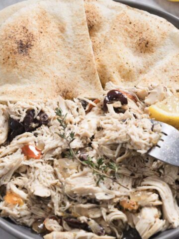 A plate featuring Greek chicken is loaded with shredded meat mixed with chopped vegetables and olives, garnished with a sprig of thyme. Two lemon slices and a fork accompany the dish, while two pieces of pita bread complete this delightful chicken recipe.