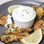 A gray plate holds several zucchini fritters, a small dish of creamy white dip with herbs, and lemon wedges. One fritter is broken to show the inside. A spoon rests in the dip.