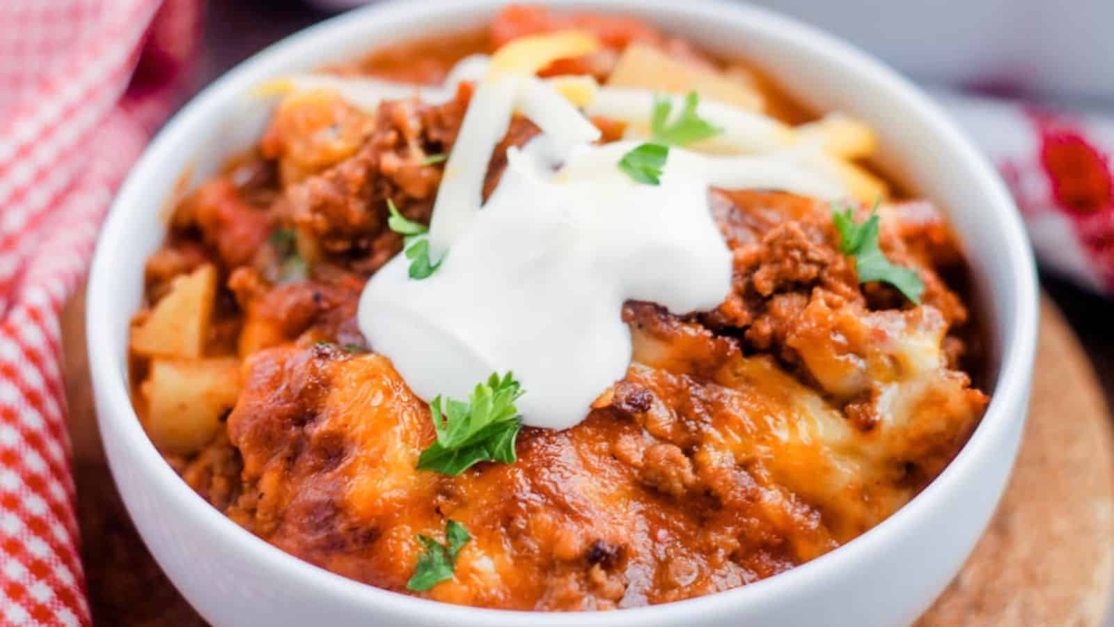 A bowl of chili topped with shredded cheese, sour cream, and parsley is perfect for easy potluck recipes. The chili boasts visible chunks of beef and beans, with a sprinkle of melted cheese on top. A red and white checkered cloth is partially visible beside the bowl, ready for sharing.