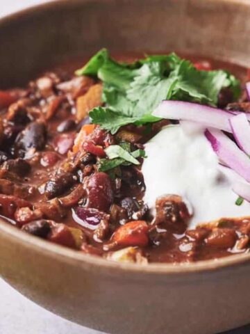 A brown ceramic bowl filled with chili, garnished with sliced red onions, a dollop of sour cream, and fresh cilantro leaves. The dish offers visible chunks of vegetables and beans. Perfect for winter, it's a recipe that has us obsessed. A sprig of fresh cilantro lies beside the bowl on the table.
