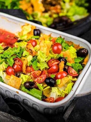 A white baking dish filled with a layered salad featuring chopped lettuce, cherry tomatoes, black olives, green onions, and tortilla strips sits atop a dark cloth. Perfect for a cozy night in,
this vibrant dish is beautifully backdropped by a blurred salad bowl.