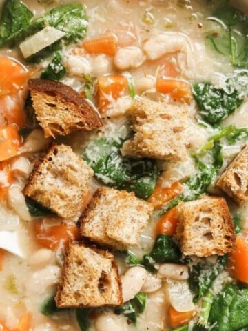 A bowl of soup featuring white beans, spinach, and carrots, topped with cubed pieces of crusty bread. A spoon rests in the bowl. The soup appears creamy and is garnished with grated cheese.