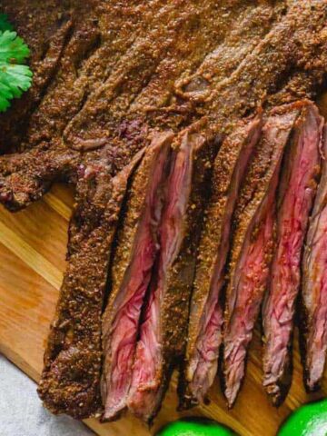 Grilled steak sliced into medium-rare strips on a wooden cutting board. Fresh cilantro and lime wedges are placed beside the steak.