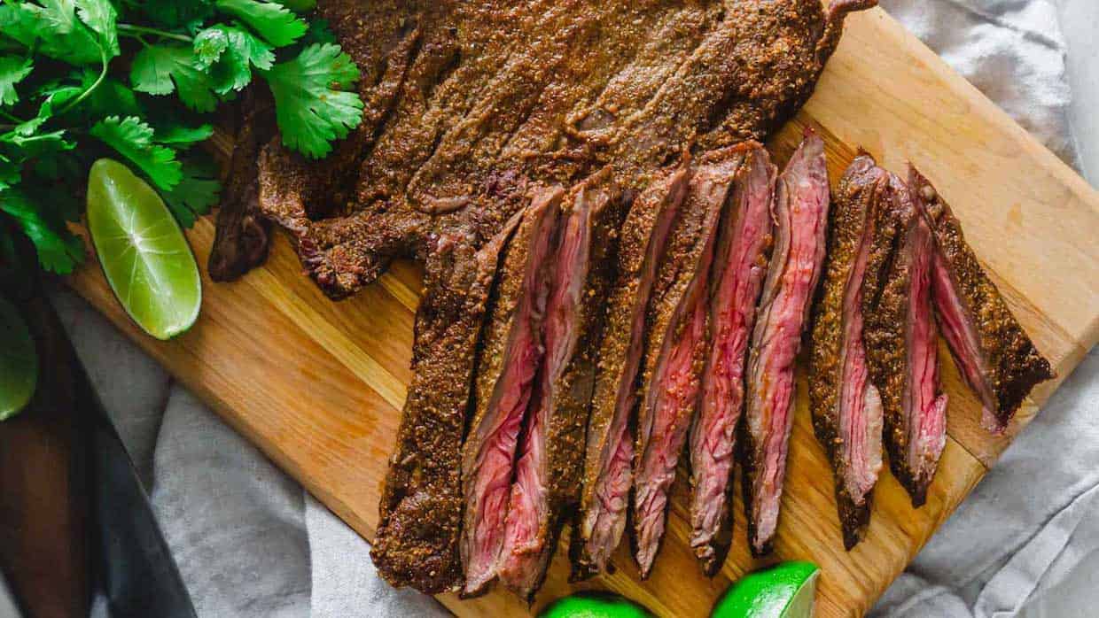 Grilled steak sliced into medium-rare strips on a wooden cutting board. Fresh cilantro and lime wedges are placed beside the steak.