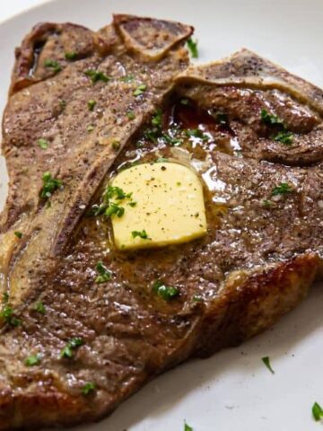 A T-bone steak on a white plate, topped with a slice of melted butter and garnished with chopped parsley. The steak appears freshly cooked with a browned exterior. A dark napkin is partially visible in the background.