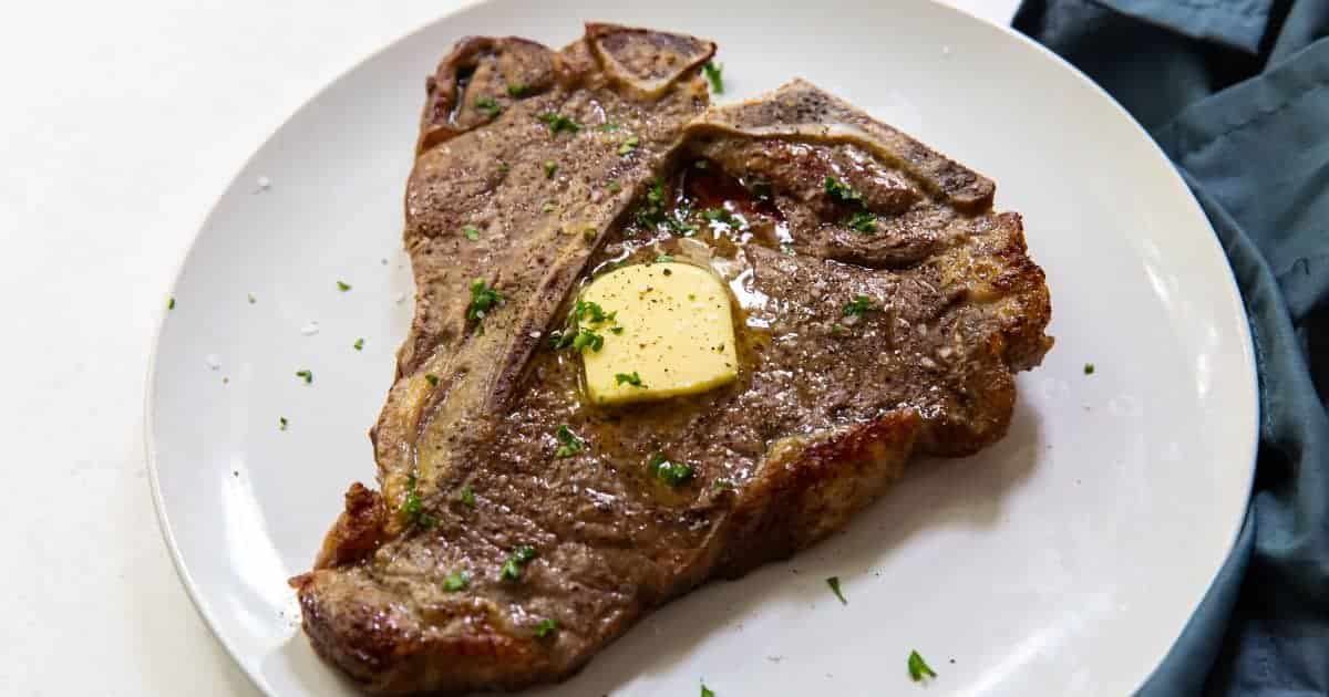 A T-bone steak on a white plate, topped with a slice of melted butter and garnished with chopped parsley. The steak appears freshly cooked with a browned exterior. A dark napkin is partially visible in the background.