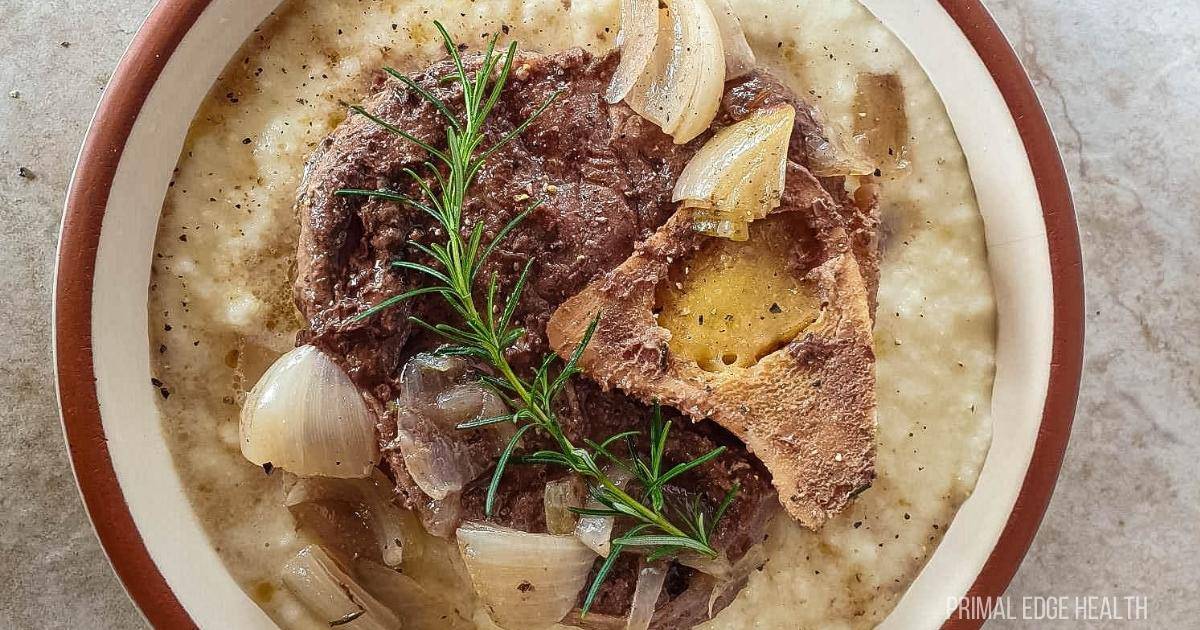 A stress-free dinner idea, this bowl of creamy porridge is topped with bone-in meat, cooked onions, and garnished with fresh rosemary sprigs. Seasoned with visible black pepper, the dish rests invitingly on a stone-like surface.