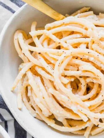 A bowl of creamy spaghetti is topped with a sprinkle of black pepper. The pasta sits on a white and blue striped cloth, with a wooden object partially visible in the background.