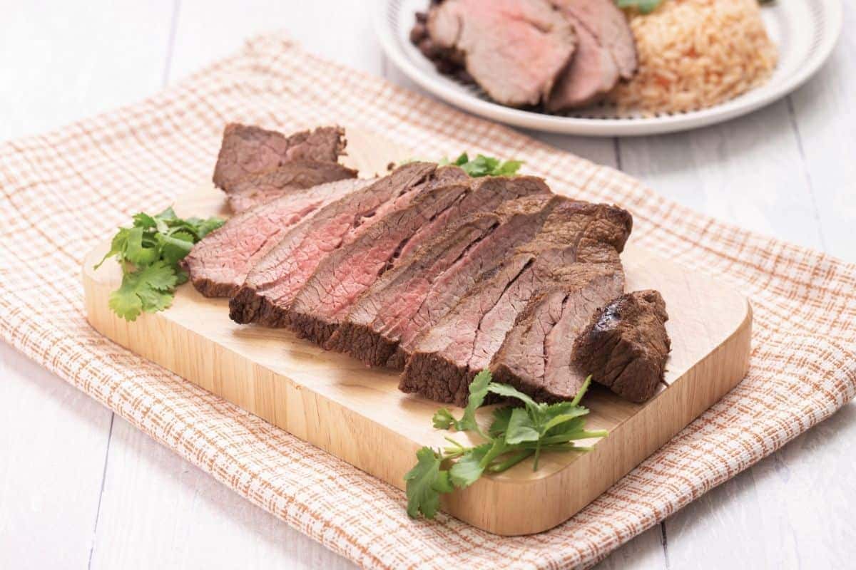 Sliced medium-rare beef steak on a wooden board, garnished with fresh herbs, placed on a checkered cloth. In the background, showcasing an effortless oven meal, a plate with additional beef slices and a serving of rice is visible.