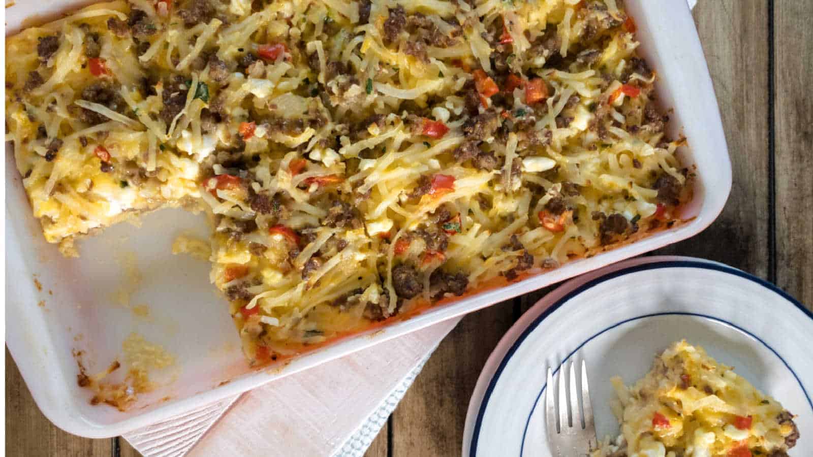 A delicious baked casserole dish filled with shredded hash browns, ground meat, and chopped red and green vegetables sits enticingly on a wooden table. This hearty casserole recipe is served in a generous portion on a white plate with a fork beside it.