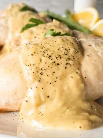 A close-up of chicken breasts topped with creamy sauce and black pepper on a white plate, showcasing how low-effort recipes can still be gourmet. Two lemon wedges and parsley garnish add the perfect finishing touch in the background.