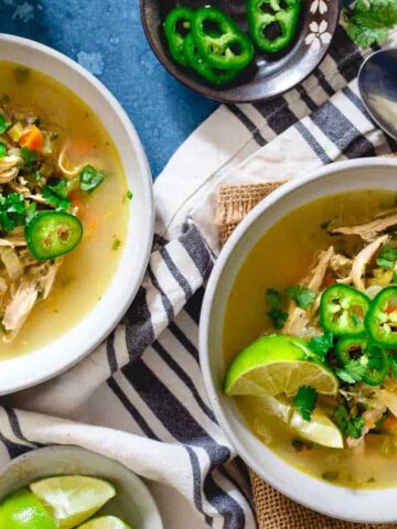 Two bowls of chicken soup garnished with lime wedges, sliced jalapeños, and cilantro are placed on a cloth next to a spoon. Additional lime and jalapeños are in small bowls nearby. The background is a blue textured surface.
