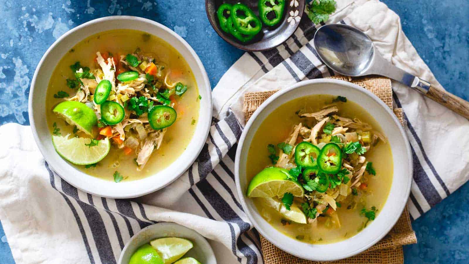 Two bowls of chicken soup garnished with lime wedges, sliced jalape&ntilde;os, and cilantro are placed on a cloth next to a spoon. Additional lime and jalape&ntilde;os are in small bowls nearby. The background is a blue textured surface.