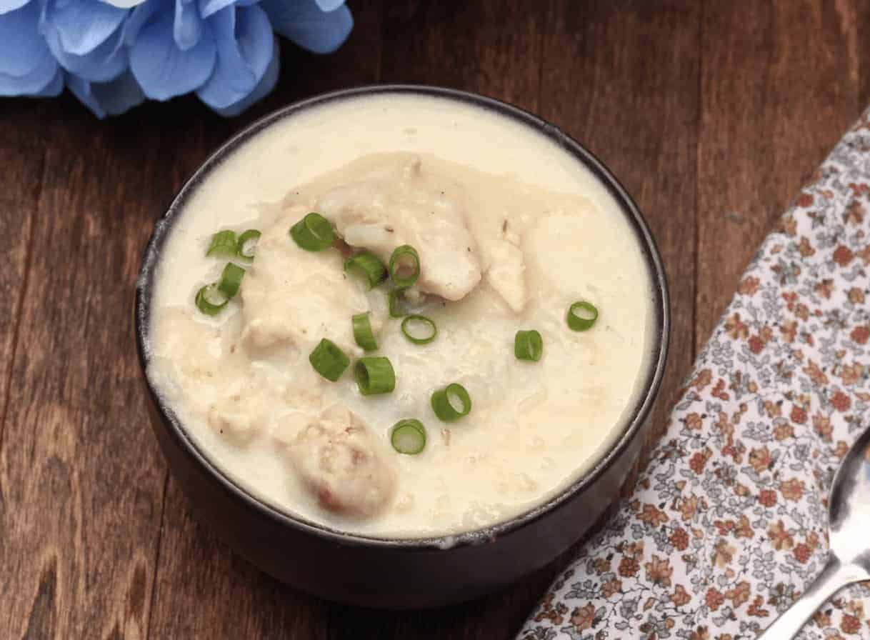 A bowl of creamy, toasty soup with chunks of chicken and sliced green onions on top rests on a wooden table. A flowery napkin and spoon are positioned to the side, while a blue flower peeks in the corner&mdash;the perfect winter meal.