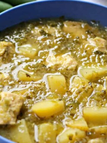 A blue bowl brimming with hearty soup, showcasing chunks of meat and potatoes in a green broth—a perfect winter meal. Nearby, a spoon and folded white cloth rest on the table, while a green chili pepper peeks from the background, making it an ideal snow day treat.