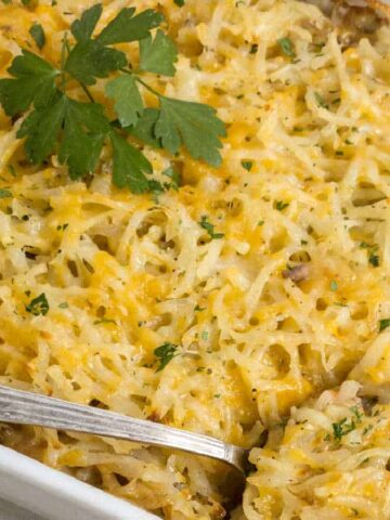 A rectangular dish showcases a delectable casserole topped with melted cheese, garnished with a sprig of parsley. A silver serving spoon rests in the dish, and the edges are enticingly browned—a perfect addition to your favorite casserole recipes.