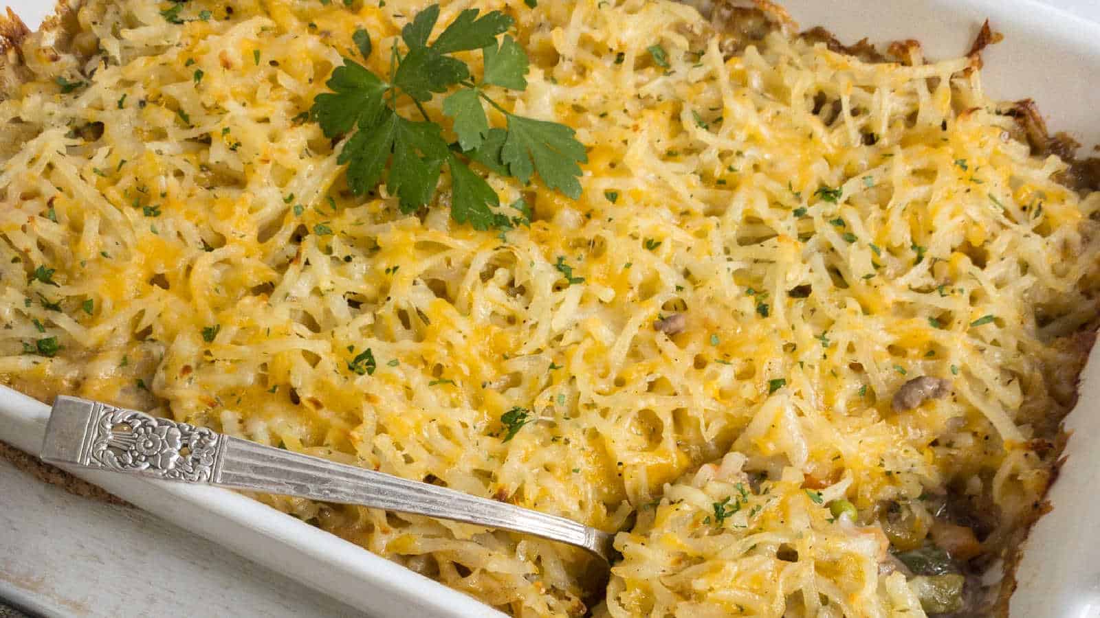 A rectangular dish showcases a delectable casserole topped with melted cheese, garnished with a sprig of parsley. A silver serving spoon rests in the dish, and the edges are enticingly browned&mdash;a perfect addition to your favorite casserole recipes.