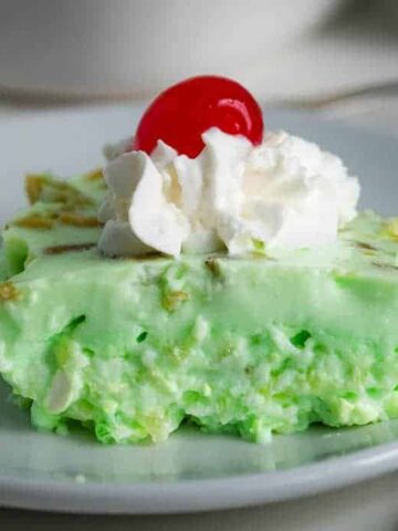 A slice of light green dessert sits on a white plate. It is topped with a dollop of whipped cream and a red cherry. The dessert has a creamy texture with small bits mixed in. A green and white cloth is partially visible in the background.