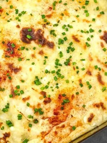 A baking tray with baked mashed potatoes topped with melted cheese and sprinkled with chopped chives sits ready for a quick cooking session. A serving spoon rests in the upper left corner, and a portion has been removed. A floral-patterned cloth is partially visible on the right.