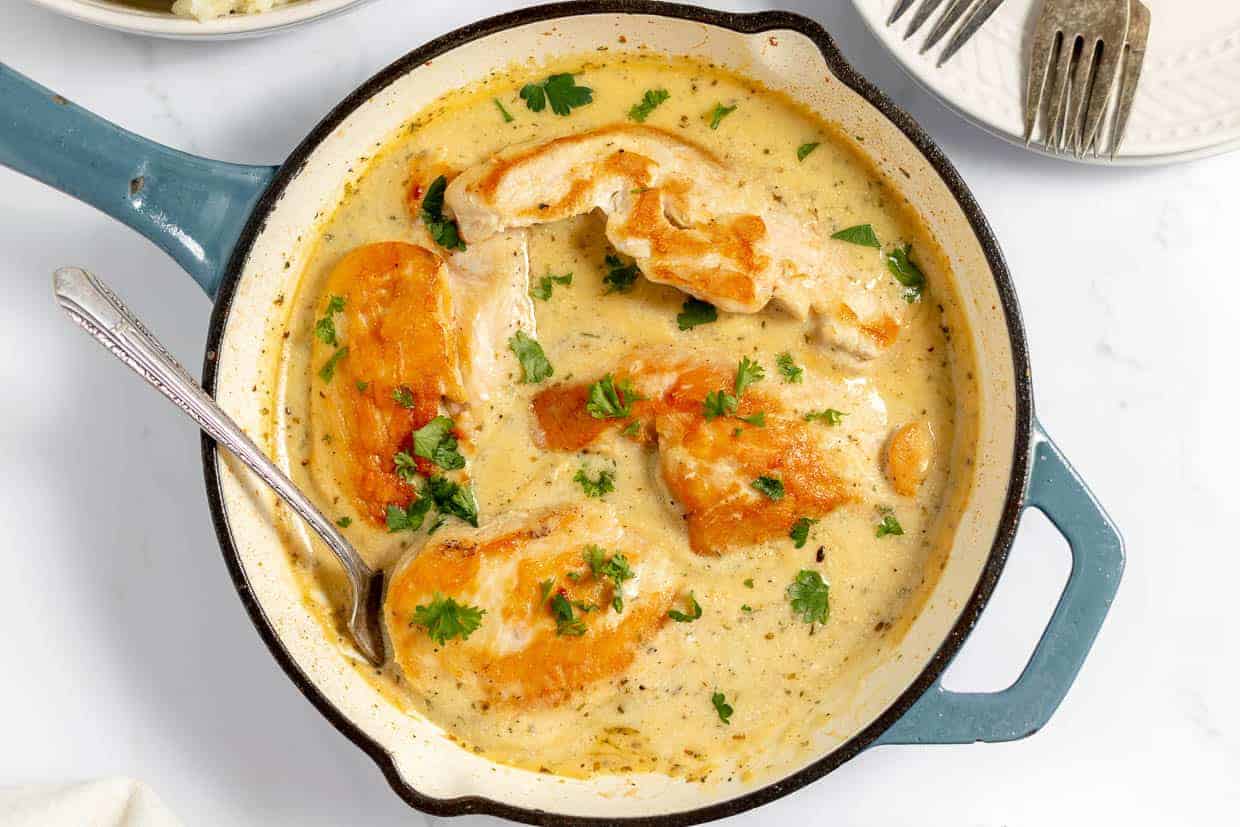 A skillet of creamy almond chicken garnished with fresh parsley offers the ultimate comfort food to keep winter blues at bay. A metal spoon rests invitingly in the dish, while two empty white plates with forks sit eagerly in the background.