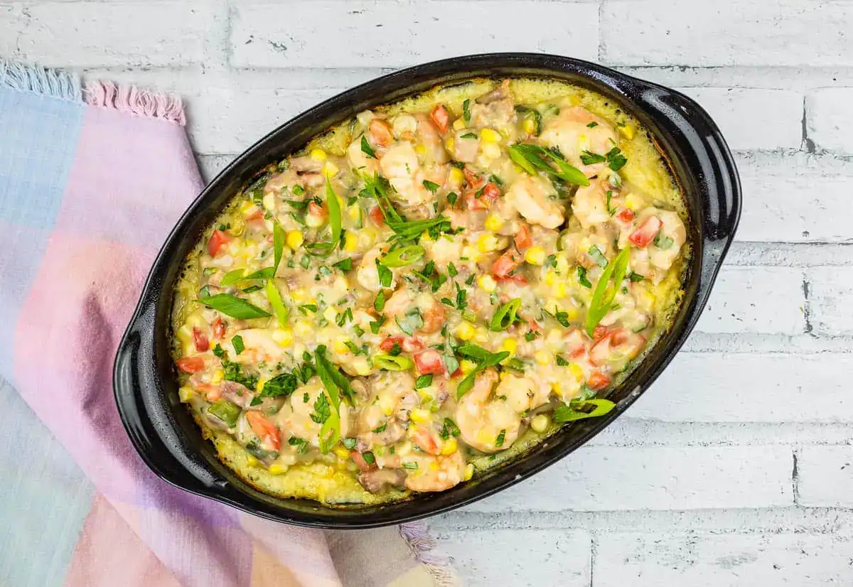 This comforting casserole features a delightful blend of shrimp, corn, diced tomatoes, and green herbs. Perfect as a winter recipe, it is elegantly presented in a black oval dish on a white brick surface with a multicolored cloth nearby.