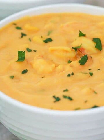 A white bowl filled with creamy cheddar cheese soup, garnished with chopped parsley. A spoon is placed nearby on a textured surface.
