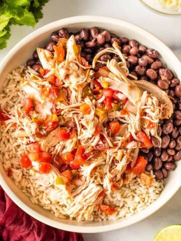 A bowl of shredded chicken, diced tomatoes, black beans, and rice is garnished with cilantro, offering a filling meal to toast any winter day. A red napkin with a fork is on the left, with lime halves and small bowls of sour cream nearby.