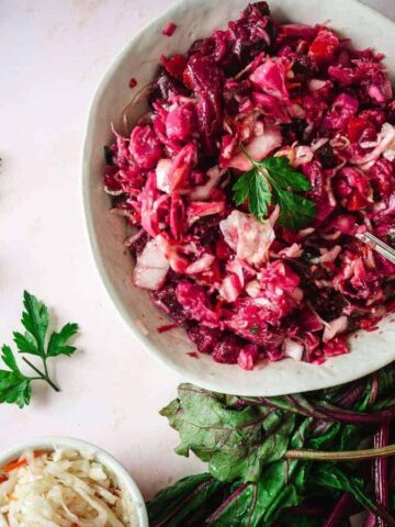 A person's hand holds a fork, digging into a bowl of red cabbage and beet salad. Fresh parsley is scattered on the surface, with additional parsley and beet greens nearby. An extra small bowl of salad is on the side.