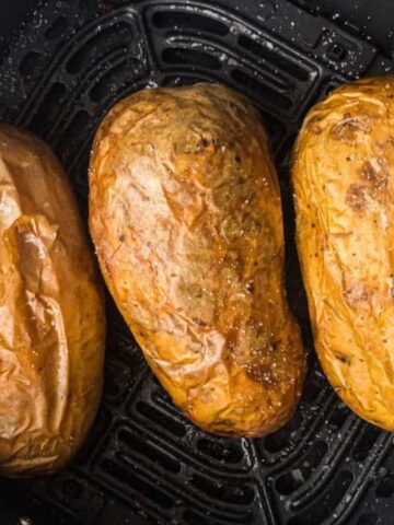Three baked potatoes are placed side by side inside an air fryer basket. The potatoes have a seasoned, golden-brown skin, indicating they are thoroughly cooked. The air fryer basket is black with a textured grate at the bottom.