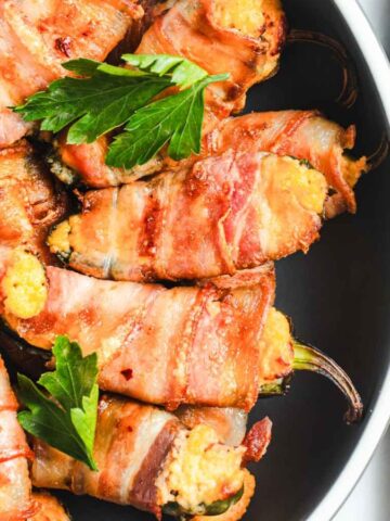 Plate of bacon-wrapped peppers filled with cheese, garnished with parsley, next to a grated cheese block and grater on a wooden board.