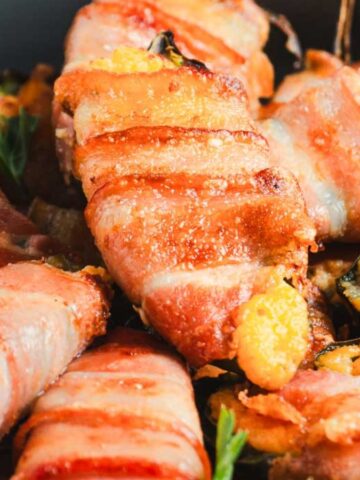 Bacon-wrapped jalapeño poppers are arranged on a plate, garnished with green parsley. Each popper is covered in crispy bacon, and visible yellow cheese is slightly melted, oozing from the ends.