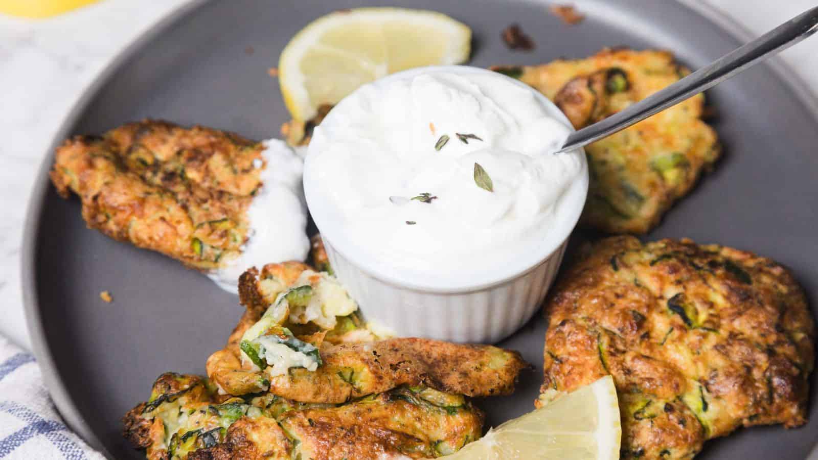 A hand dips a fried zucchini fritter into a small bowl of white cream sauce on a gray plate. Other fritters and lemon wedges are arranged around the bowl on a marble surface.
