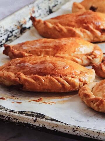 A baking tray with eight golden-brown pasties arranged in two rows on parchment paper. The pasties have crimped edges and are evenly baked, with a flaky, glossy crust. The tray shows signs of use, with a slightly textured and worn appearance.