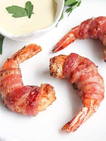 Three bacon-wrapped shrimp are arranged on a white plate with sprigs of parsley. A small dish of creamy sauce is placed at the top.