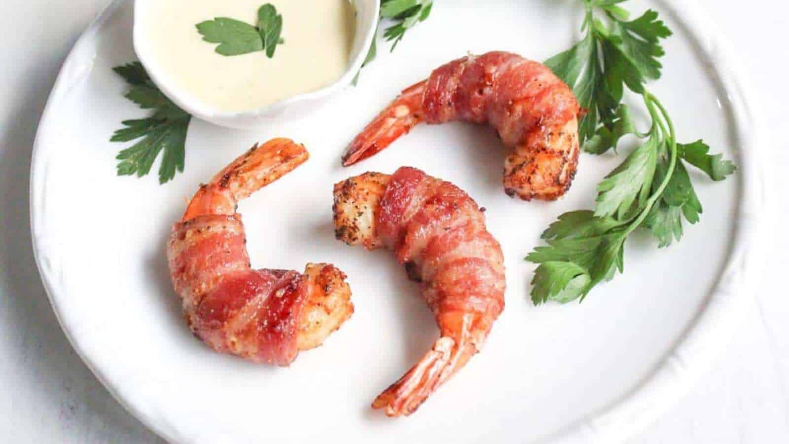 Three bacon-wrapped shrimp are arranged on a white plate with sprigs of parsley. A small dish of creamy sauce is placed at the top.