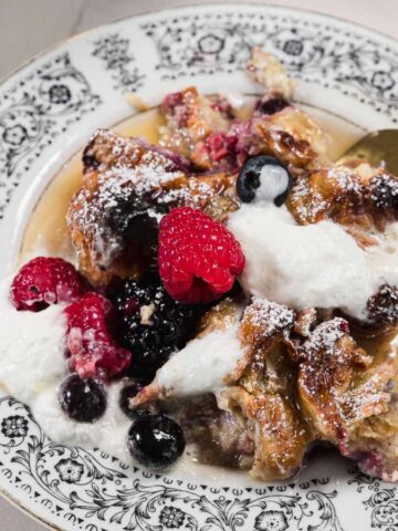 A dessert on a decorative plate with a golden fork. The dish features bread pudding topped with whipped cream and assorted berries, including raspberries, blueberries, and blackberries, sprinkled with powdered sugar.
