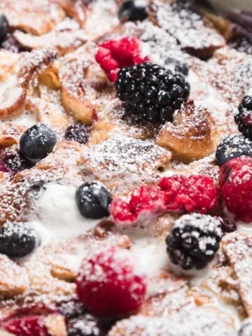 A baked dessert with toasted bread pieces, topped with fresh raspberries, blackberries, and blueberries. The dish is dusted with powdered sugar, and a light drizzle of cream is visible on top.