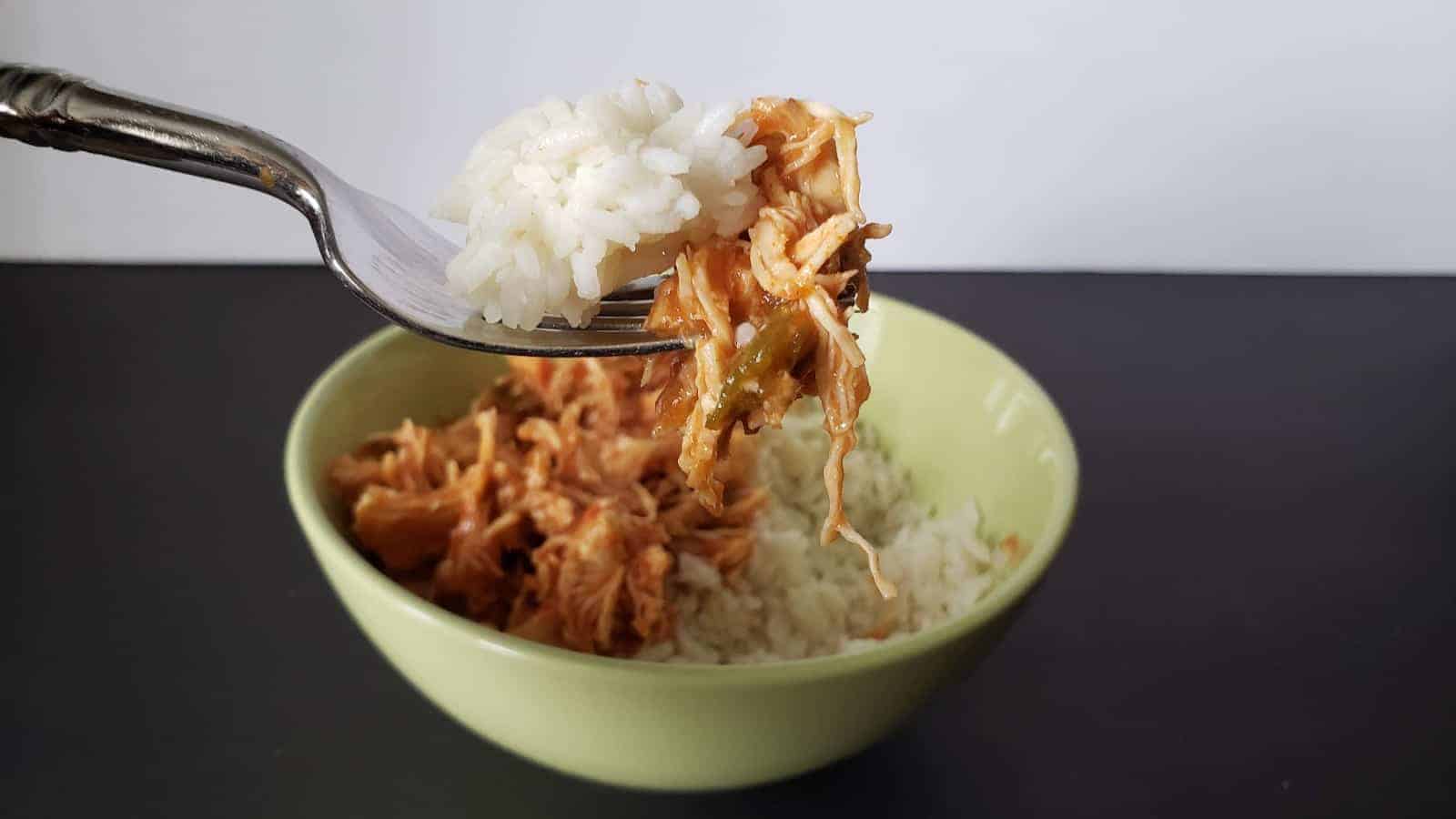 A fork with a bite of white rice and shredded meat is held above a green bowl. The bowl contains a serving of white rice and shredded meat mixed with sauce. The background is black and white.