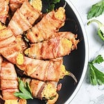 A plate filled with flavorful air fryer jalapeño poppers, stuffed with a rich cheese mixture and wrapped in crispy bacon. The poppers are beautifully arranged on a round, dark plate, accented by fresh parsley leaves for garnish.