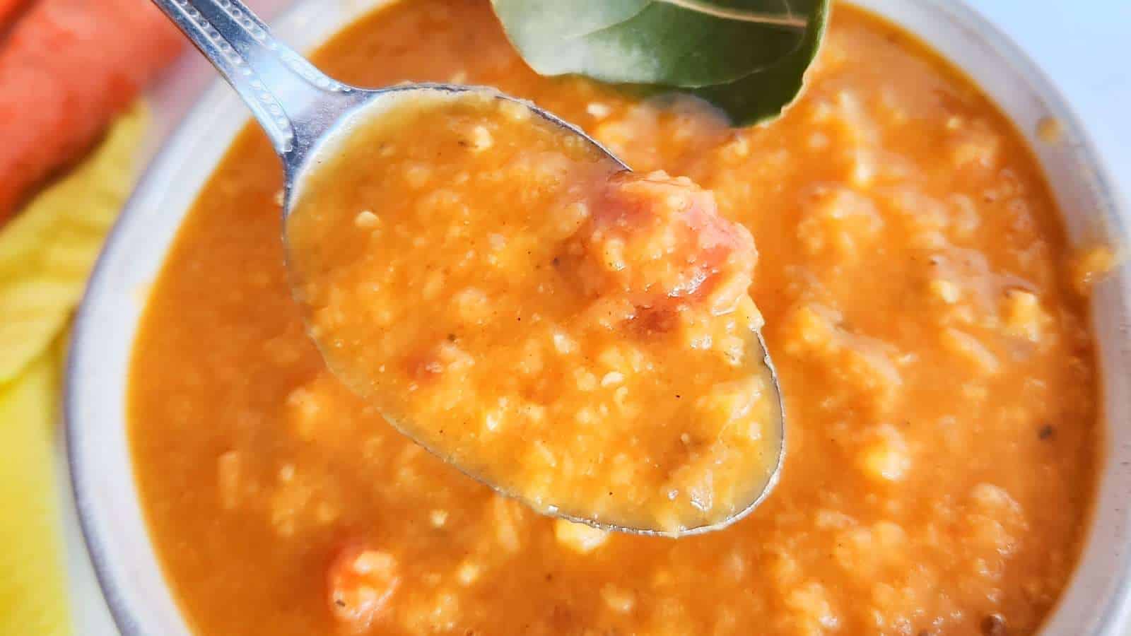 A close-up of a spoonful of thick, orange lentil soup with visible chunks of tomato, held over a bowl filled with the same soup. A bay leaf is visible on the surface of the soup in the bowl.