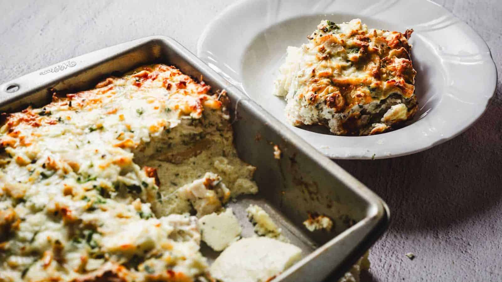A baked casserole dish filled with a cheesy, golden-brown topping is being scooped with a spatula. The dish appears to contain layers of ingredients beneath the melted cheese, with herbs sprinkled on top for garnish.