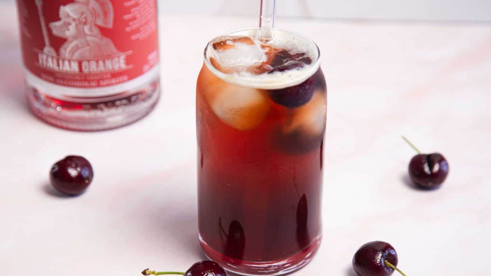 A tall, clear glass filled with a dark red drink, ice cubes, and garnished with cherries. A clear straw is inserted into the drink. Surrounding the glass are fresh cherries, with a bottle labeled 