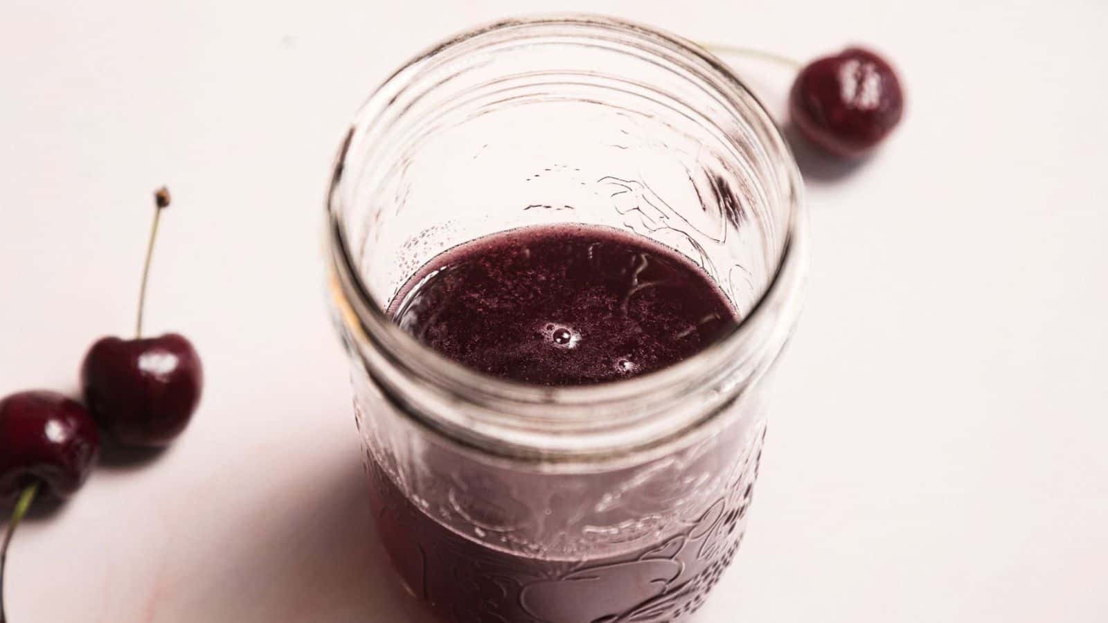 A glass jar filled with a dark red liquid sits on a light surface, viewed from above. Three fresh cherries with stems are placed nearby.