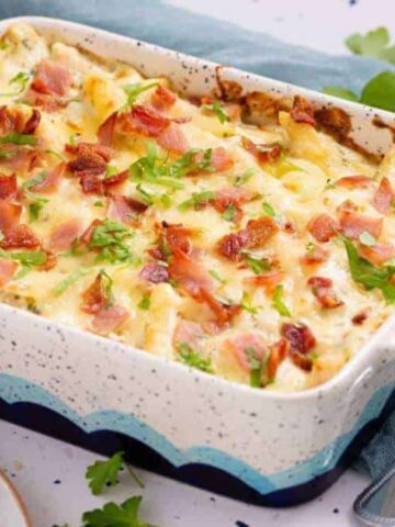 A baked pasta casserole in a speckled rectangular dish, topped with melted cheese, diced ham, and chopped parsley. A silver serving spoon and a plate with a fork are placed nearby on a light-colored tablecloth.