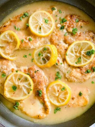 A pan featuring chicken breasts cooked in a creamy lemon sauce, garnished with lemon slices and parsley. The dish is placed on a light-colored surface with a wooden handle of the pan visible.