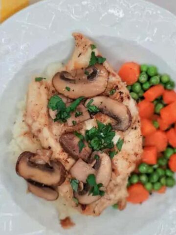 A white plate with grilled chicken topped with mushrooms and herbs, served over mashed potatoes. It is accompanied by a side of peas and carrots. A yellow checkered cloth and utensils are next to the plate.
